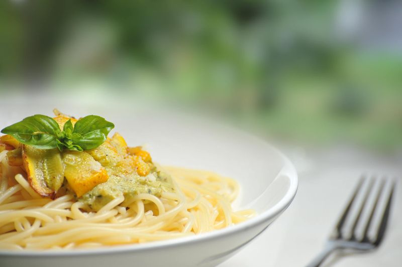 White bowl of spaghetti with green vegetable sauce, roasted squash, and fresh basil on a sunlit surface
